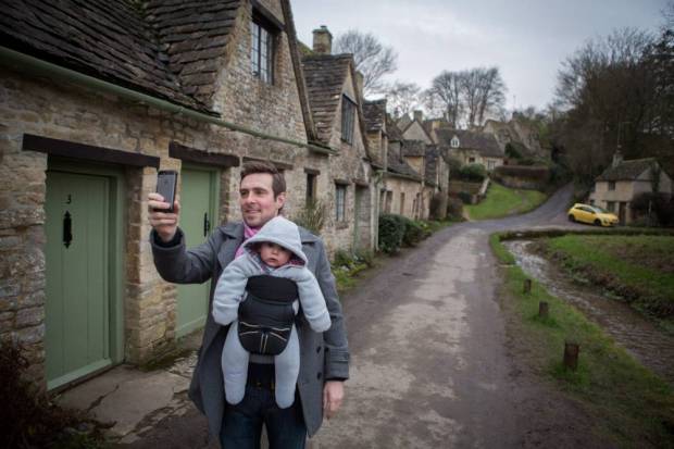 Adam Petrie, 36, takes a selfie with his son Fred, 4 months old, at Arlington Row in Bibury, Cotswolds, where a yellow car has been the cause of controversy for spoiling the view. See SWNS story SWYELLOW; A bright yellow car is photo-bombing England's most picturesque view. Tourists' picture-postcard shot of the Cotswold's Arlington Row are being ruined by "ugly" car - which is appearing in photographs all over the world. The 14th century cottages, owned by the National Trust, are the most photographed cottages in Britain and attract swarms of visitors every year. The car's owner, Peter Maddox, 82, said there was nothing he could do about the banana yellow car.