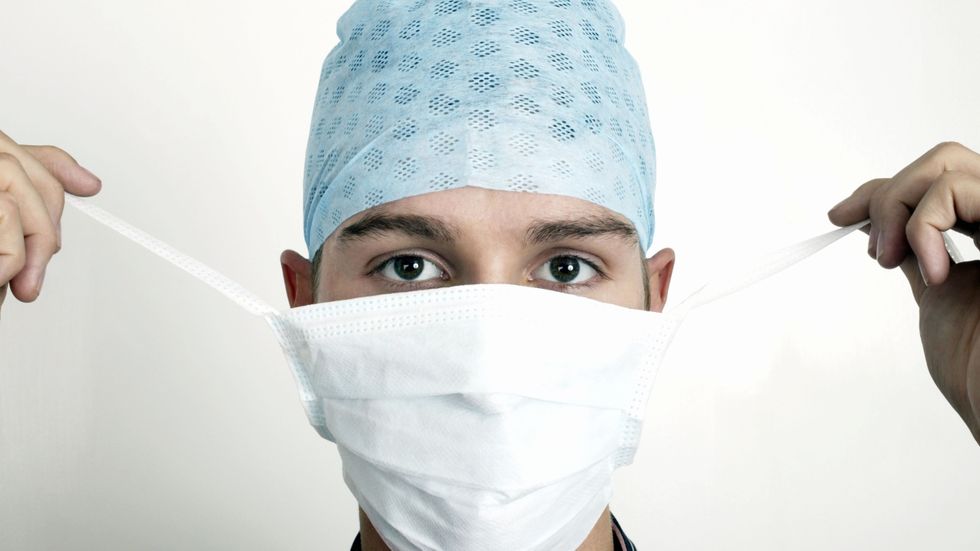 Close up of face of young doctor, wearing blue surgical cap and tying up white hygiene mask in preparation for surgery (Photo by Universal Images Group via Getty Images)